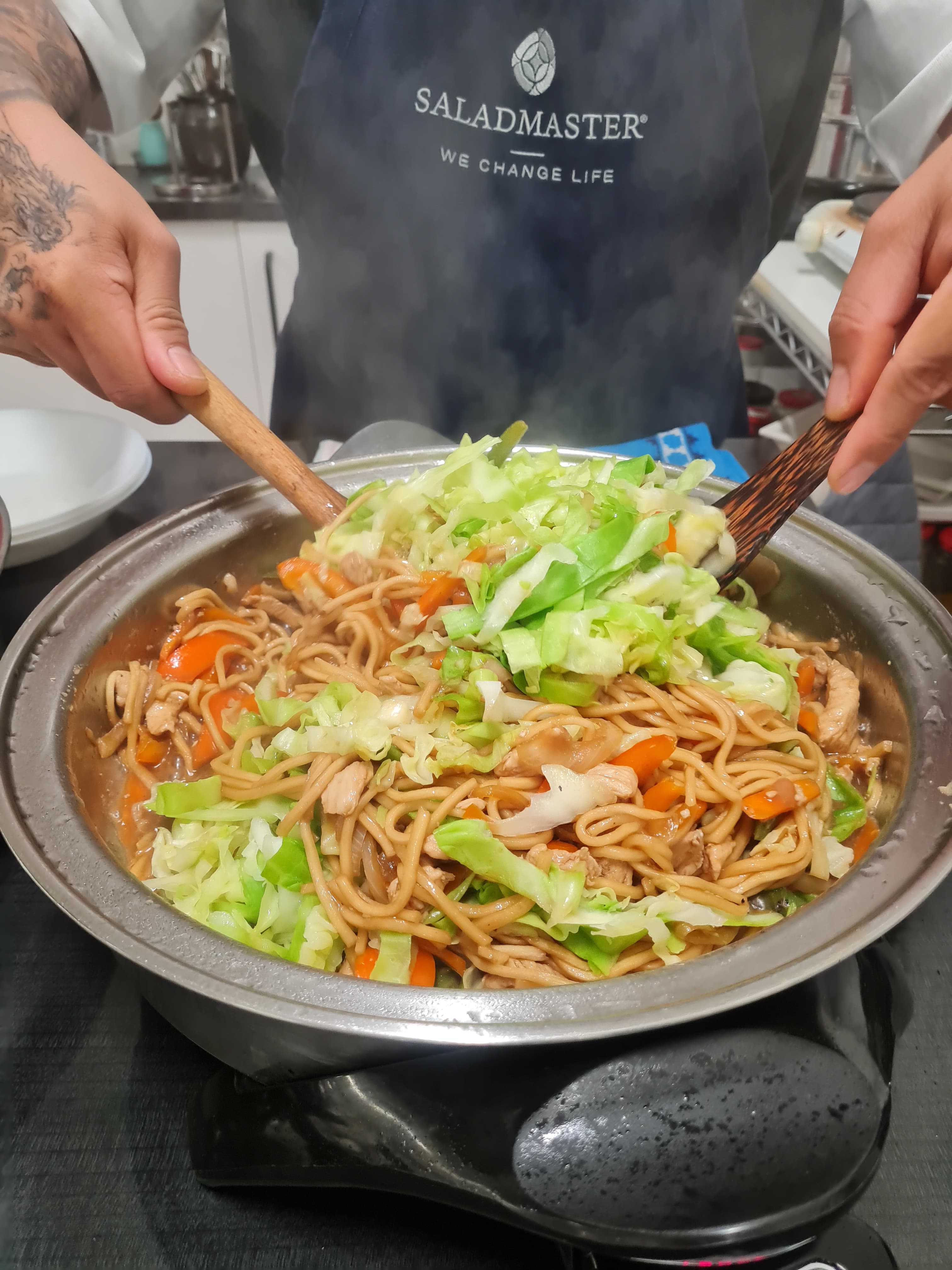 Noodles being tossed in a steaming Saladmaster pan at a dinner demo