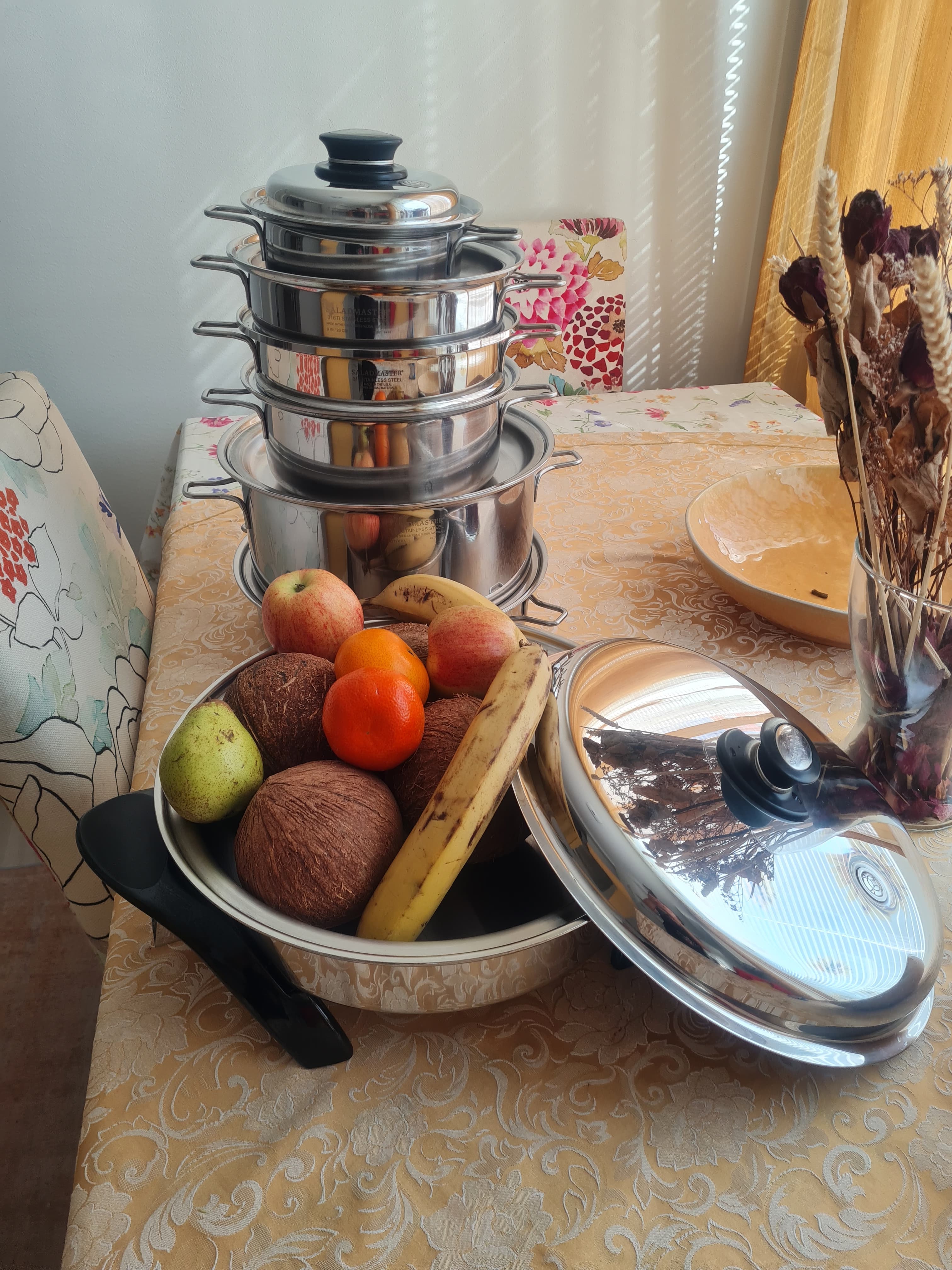A stacked Saladmaster cookware set on the kitchen table beside a pan of fresh fruit